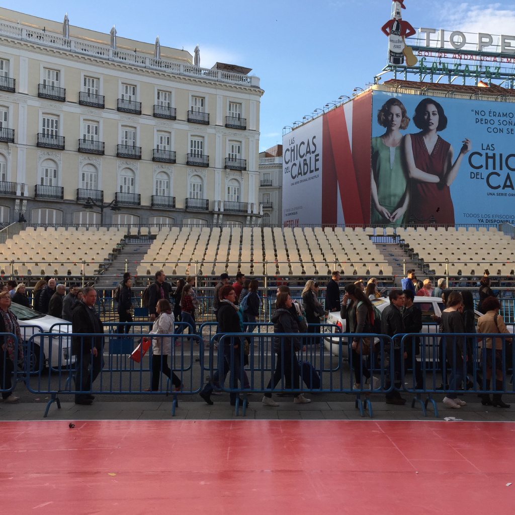 Multitud caminando frente a cartel publicitario en plaza.